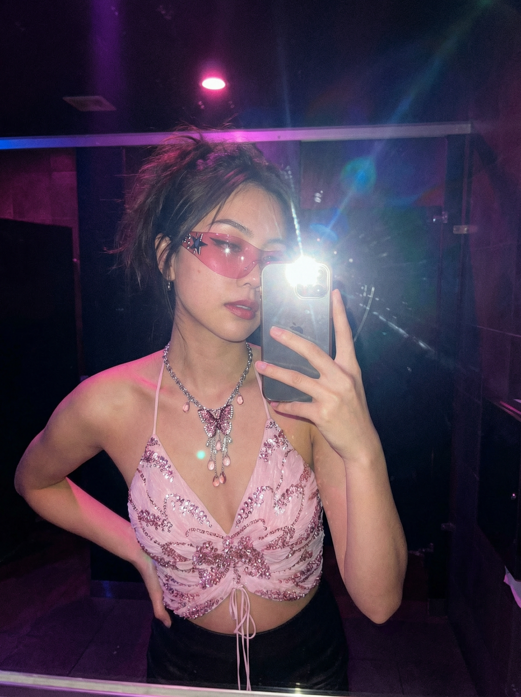 Model wearing the Petal Drip Y2K pink butterfly pendant necklace with a pink sequined top and sunglasses in a dimly lit room