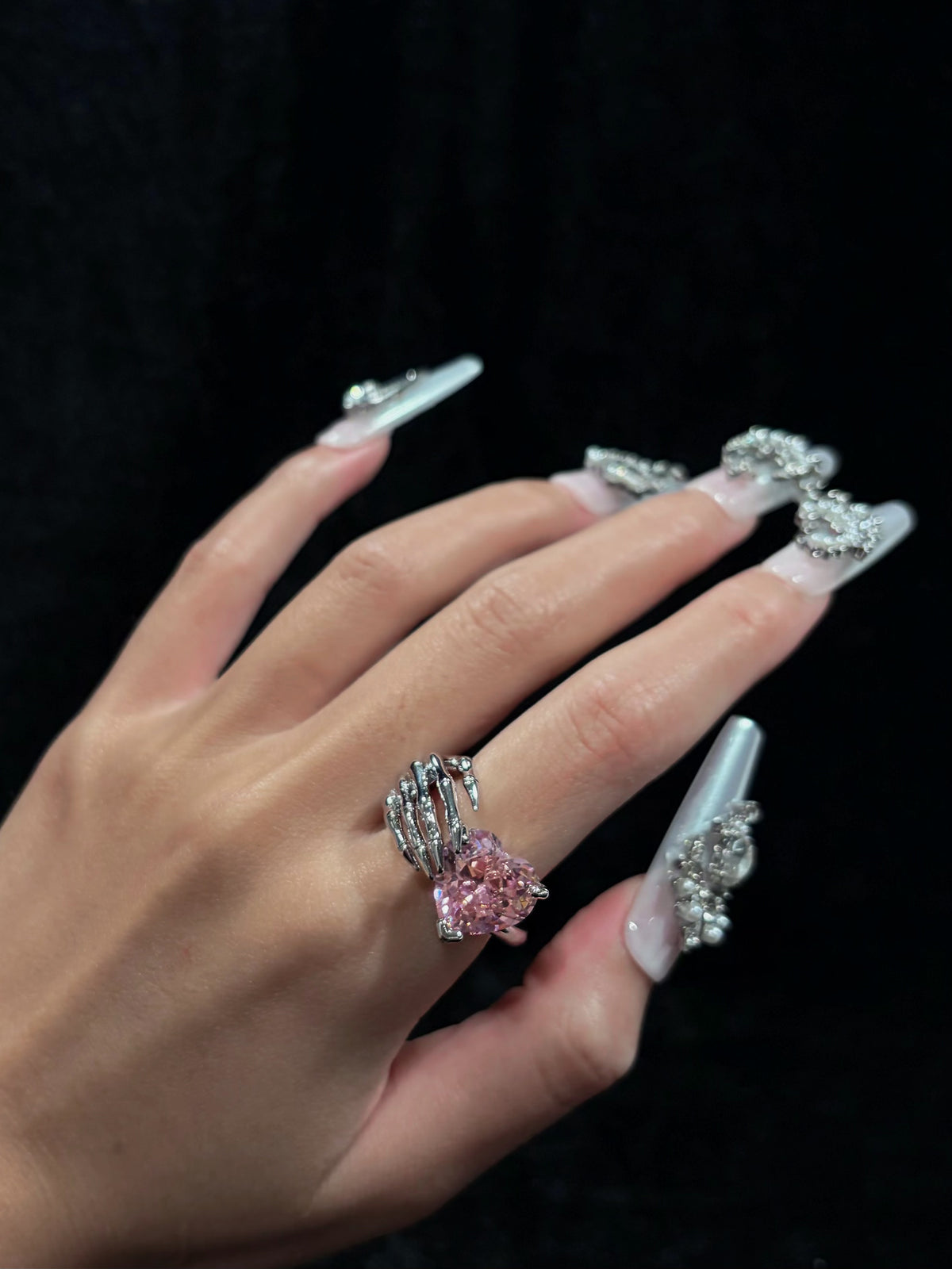 Hand wearing the Skeleton Heart Y2K sparkling adjustable pink crystal ring with a sculpted skeleton hand design against a dark background